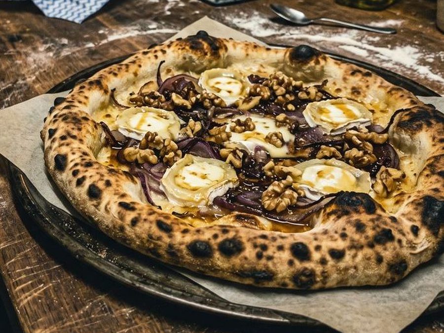 Photo prise au téléphone dans une arrière-cuisine sombre : pizza chèvre, oignons rouges, noix et miel, posée sur un plan de travail en bois